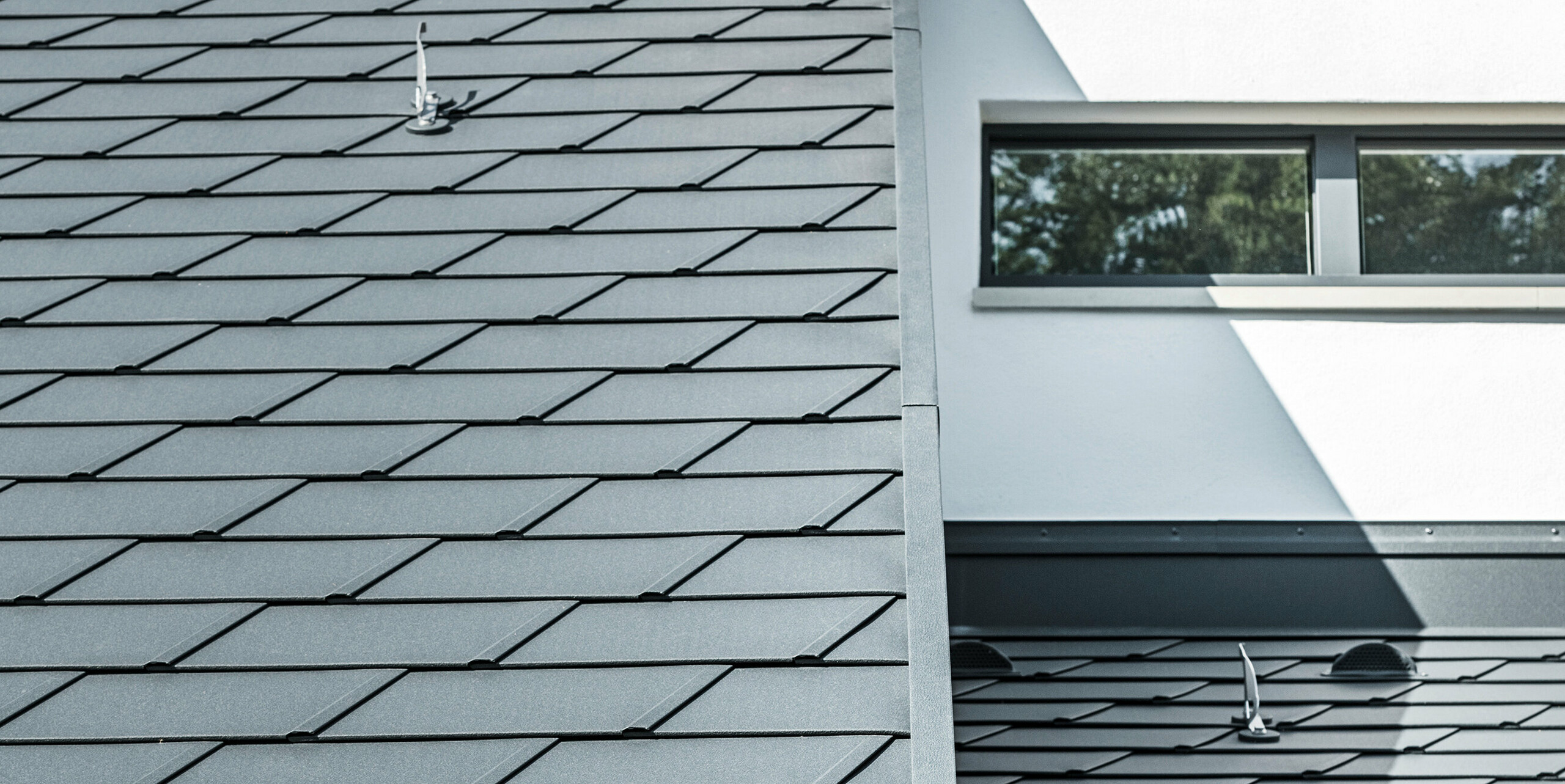 Detailní pohled na různé úrovně střechy rodinného domu v Dortmundu. Falcovaný šindel PREFA DS.19 a střešní odvodňovací systém PREFA v barvě P.10 antracitové a také bezpečnostní střešní háky PREFA.