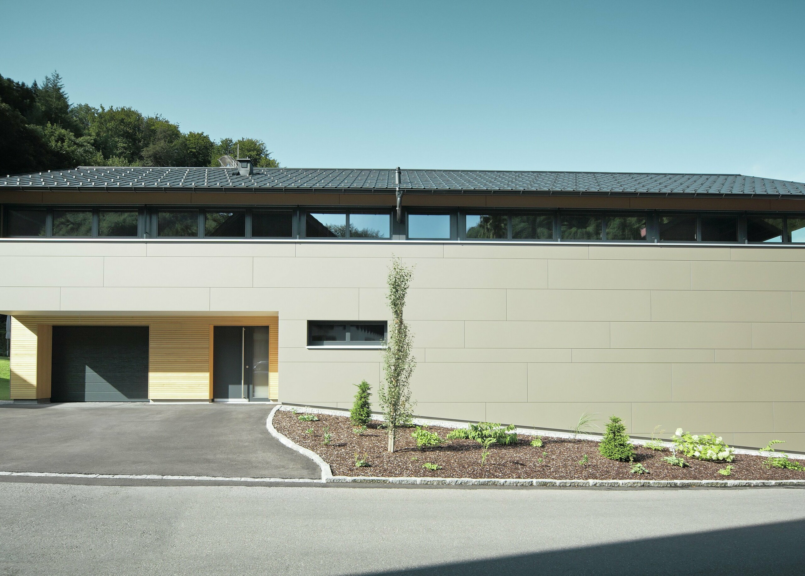 Das Einfamilienhaus in Hohenems präsentiert sich in einer modernen und stilvollen Fassade aus PREFA Aluminium Verbundplatten in Bronzemetallic, mit einem darüberliegenden Dach aus PREFA Dachplatten in P.10 Anthrazit. Die klare Linienführung und das durchdachte Farbkonzept des Designs integrieren das Gebäude nahtlos in die natürliche Umgebung. Die Gestaltung zeigt die Kombination aus Funktionalität und Ästhetik, die PREFA Produkte für zeitgemäße Wohnarchitektur bieten.