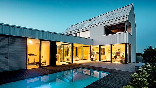 Obrázek ukazuje moderní rodinný dům ve Freistadtu s výraznou fasádou PREFALZ v čistě bílé barvě. Precizně skládaná hliníková fasáda dodává budově čistou, geometrickou eleganci, která ladí s velkorysými okny a plochou střechou. Použití PREFALZ zajišťuje nejen estetickou stálost, ale také vysokou odolnost vůči povětrnostním vlivům a trvanlivost. Otevřený interiér s výhledem na bazén je dokonale doplněn čistými liniemi a minimalistickým designem, díky čemuž je dům moderním a udržitelným řešením pro bydlení.