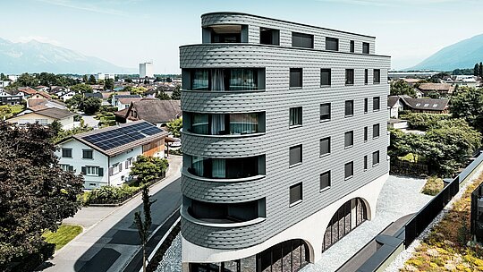 Letecký pohled na Bernerhaus v Buchsu s jeho výrazným trojúhelníkovým tvarem a jemně zaoblenými balkony. Fasáda, obložená fasádními šindeli PREFA v barvě P.10 světle šedé, obaluje budovu jako šupinovitý hliníkový plášť a vytváří živou hru světla a stínu. Nad masivním betonovým základem s elegantními arkádami působí lehká, povětrnostním vlivům odolná hliníková fasáda PREFA mimořádně precizně, trvanlivě a nenáročně na údržbu – moderní poutač uprostřed alpské městské krajiny.