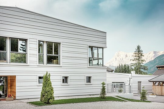 Detailní pohled na kasárna „Tempesti“ s přesně instalovanými hliníkovými šáry PREFALZ v barvě P.10 prefa bílé. Čisté linie horizontálně instalovaného obkladu se stojatou drážkou zdůrazňují moderní design fasády. Světlý povrch odráží sluneční světlo a optimalizuje mikroklima. V pozadí se tyčí impozantní jihotyrolské hory – místo, kde se technologie setkává s přírodou na nejvyšší úrovni.