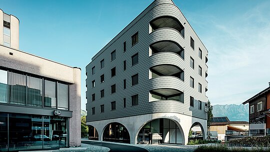 Tento moderní dům v bernském stylu v Buchsu zaujme svým výrazným, zaobleným, trojúhelníkovým tvarem a souvislou fasádou z PREFA fasádních šindelů v barvě P.10 světle šedá. Šupinovitý hliníkový povrch zdůrazňuje měkké hrany a hluboké křivky balkonů, zatímco masivní betonový základ s elegantními arkádami vytváří podmanivý kontrast. Lehká, povětrnostním vlivům odolná a trvanlivá hliníková fasáda PREFA, zasazená do alpského prostředí uprostřed malé nákupní ulice, se stává architektonickým lákadlem: je precizně vyrobená, vyžaduje jen malou údržbu a je ideální pro náročné křivky.