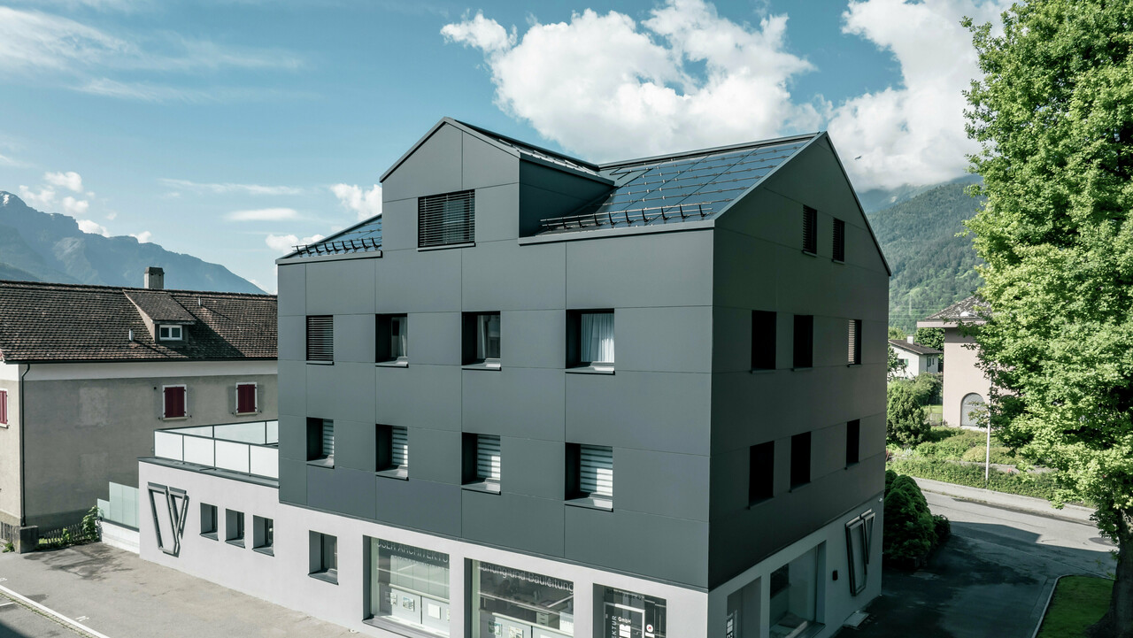 Modernisiertes Wohn- und Geschäftsgebäude nach der Fassadensanierung mit PREFABOND Aluminium Verbundplatten in Anthrazit. Die klare, elegante Linienführung der neuen Fassade verleiht dem Gebäude ein modernes Erscheinungsbild. Im Erdgeschoss befindet sich das Architekturbüro Voser. Die Umgebung mit Bäumen und Bergen rundet die harmonische Optik ab.