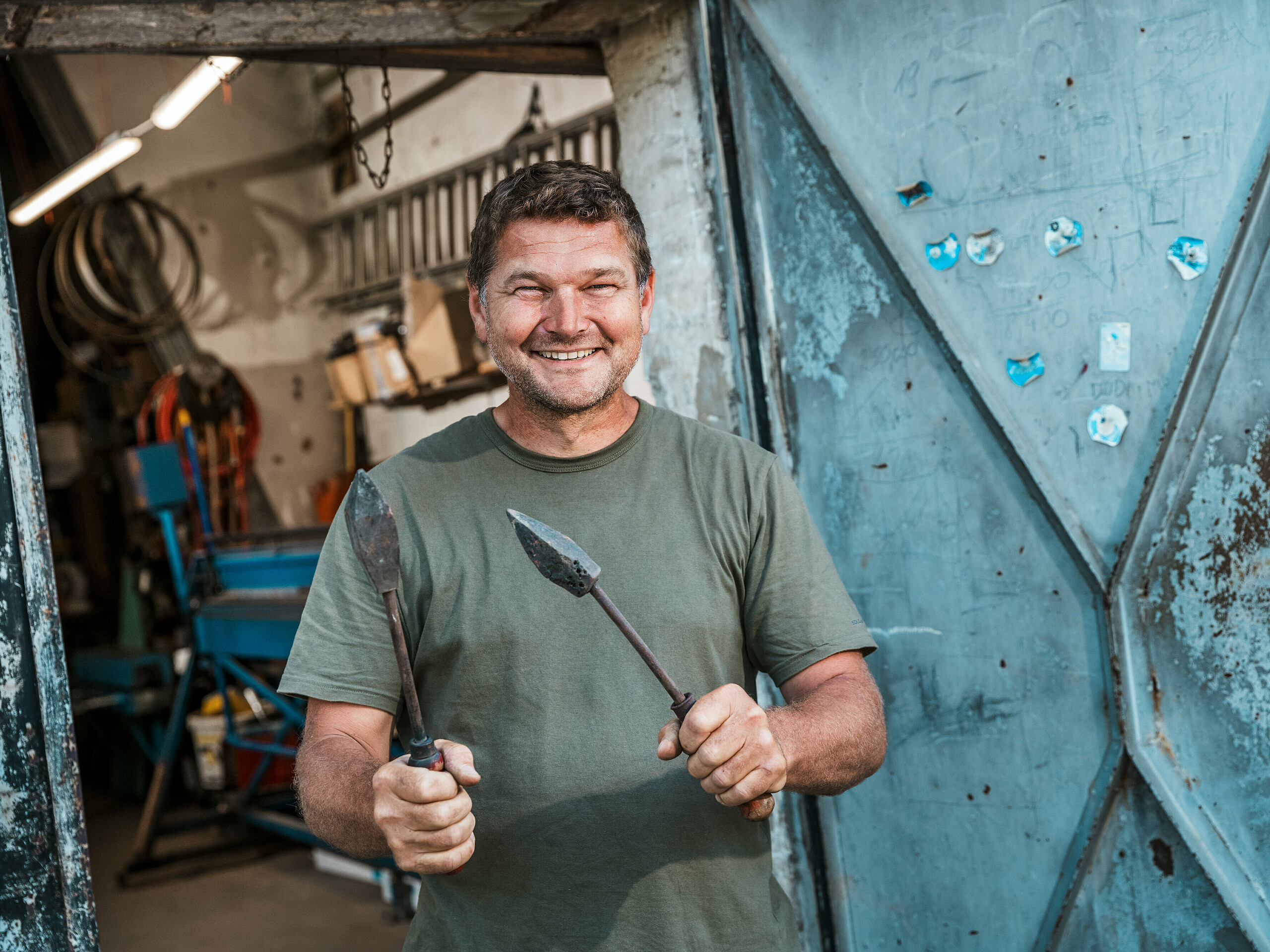 Mistr klempíř Richárd Tóth ve své dílně. Samozvaný řemeslník drží tradiční nástroje a stojí před otevřenými kovovými dveřmi. Scéna zprostředkovává autentické řemeslné zpracování a vysokou přesnost – vlastnosti, které byly klíčové při výrobě hliníkových produktů PREFA pro budapešťský bytový dům. Drsné prostředí dílny podtrhuje řemeslnou zručnost, s níž byly z materiálu PREFALZ v barvě P.10 antracitové vyrobeny fasádní prvky, parapet a obložení balkonů. Obrázek zdůrazňuje kombinaci zkušeností, materiálové odbornosti a odolných řešení PREFA.