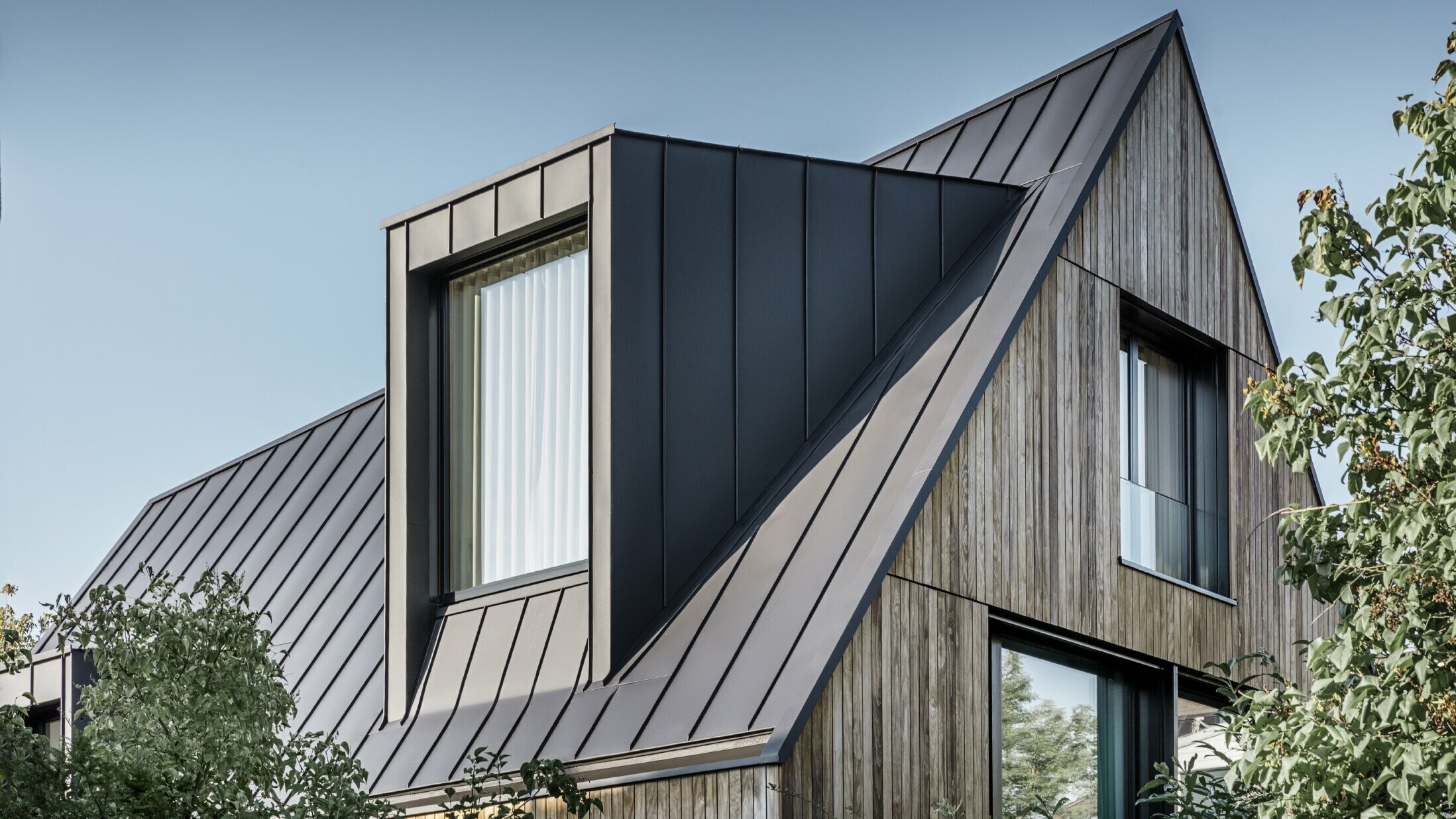 Příkrá sedlová střecha s velkým vikýřem, pokrytá PREFALZem společnosti PREFA v barvě antracitová, střecha se svitkovými plechy kombinovaná s dřevěnou fasádou