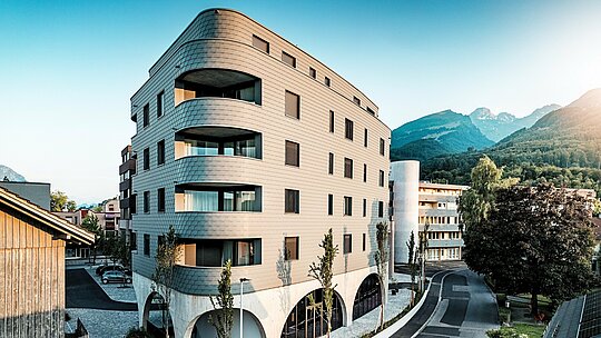 Šikmý pohled na Bernerhaus v Buchsu s jeho výrazným trojúhelníkovým tvarem a jemně zaoblenými rohy. Souvislá fasáda z fasádních šindelů PREFA v barvě P.10 světle šedé obaluje budovu jako šupinovitý hliníkový plášť a zdůrazňuje splývavé pásy balkonů. Nad masivním betonovým základem s elegantními arkádami působí obzvláště výrazně lehká, povětrnostním vlivům odolná hliníková fasáda PREFA. Přesně instalovaná, odolná a nenáročná na údržbu harmonicky zapadá do alpského prostředí a vytváří silný architektonický dojem.