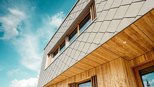 Detailní záběr na školku v Alzenau ukazuje precizní zpracování a vysoce kvalitní kombinaci bronzově zbarvených stěnových panelů PREFA (44 x 44 cm) a přírodního dřeva. Diagonální struktura hliníkové fasády vytváří výrazný šupinovitý vzor, ​​který krásně ladí s vertikálními dřevěnými žaluziemi kolem oken a precizně zpracovaným dřevěným podhledem. Záběr z nízkého úhlu zdůrazňuje přesné linie a řemeslné zpracování odvětrávaného fasádního obkladu. Tento detailní pohled podtrhuje konstrukční pevnost a trvanlivost produktů PREFA – ideálních pro udržitelné architektonické projekty, které kombinují estetiku, funkčnost a ekologickou odpovědnost na nejvyšší úrovni.