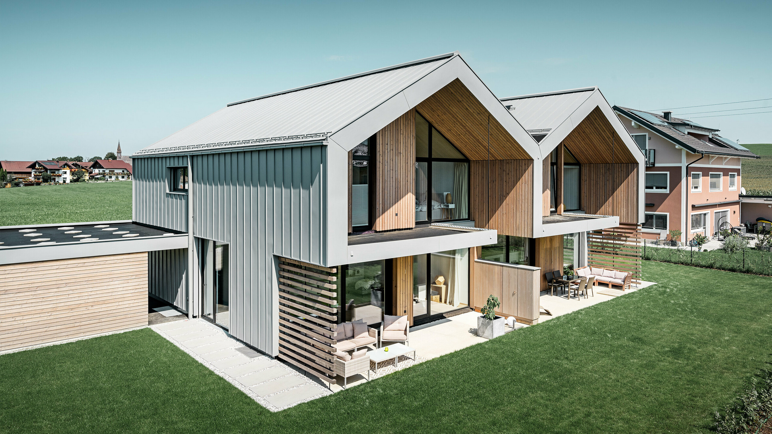 Moderní dvojdomky, jejichž střecha a fasáda jsou pokryté svitkovým plechem PREFALZ v barvě šedá patina, včetně hranatého svodu