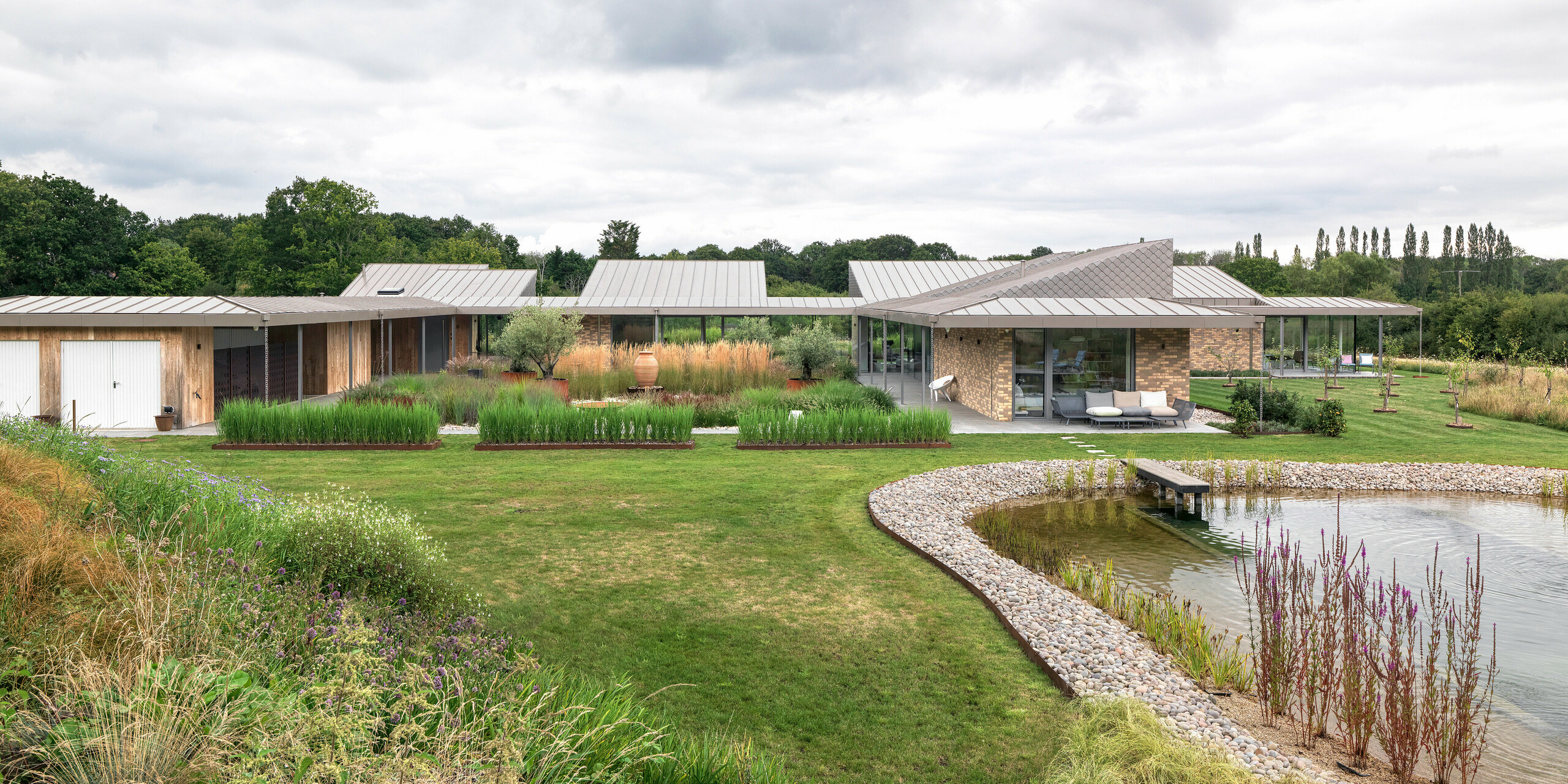 Weitwinkelansicht des architektonisch anspruchsvollen Wohnhauses in West Sussex mit großflächigem PREFALZ Aluminiumdach in P.10 Bronze und harmonisch integrierten PREFA Wandrauten in ausgewählten Fassadenbereichen. Das moderne Gebäude erstreckt sich flach und weitläufig über das Grundstück, eingebettet in eine naturnahe Bepflanzung und mit einem gestalteten Naturpool im Vordergrund. Die robusten PREFA Produkte unterstützen das nachhaltige Baukonzept und sorgen für ein einheitliches, langlebiges Erscheinungsbild.