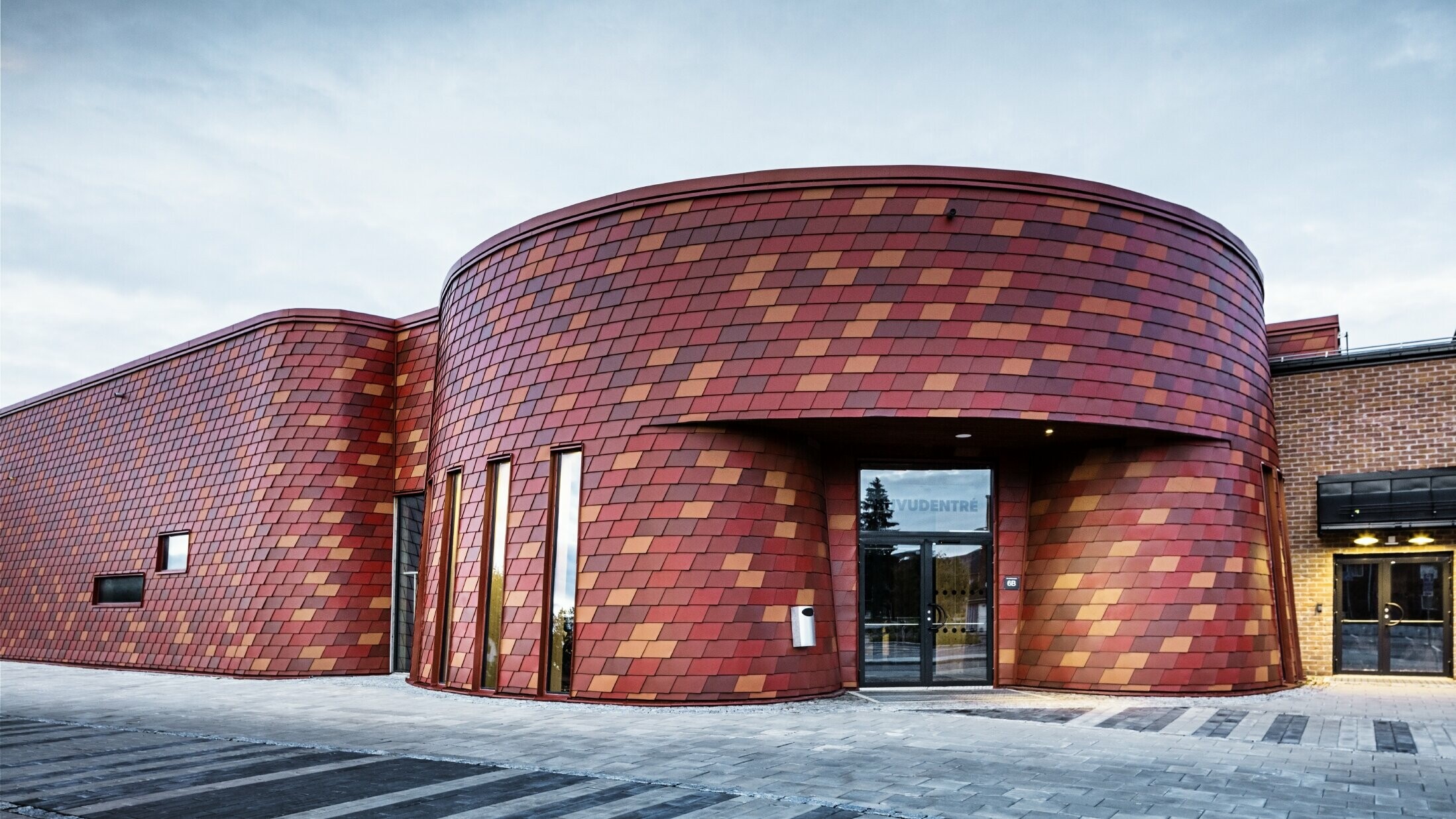 Zimní stadion s plochou střechou a zvlněnou fasádou, která je obložena PREFA fasádními hliníkovými šindeli v různých odstínech červené – tmavě červená, cihlově červená, červenohnědá, hnědočervená