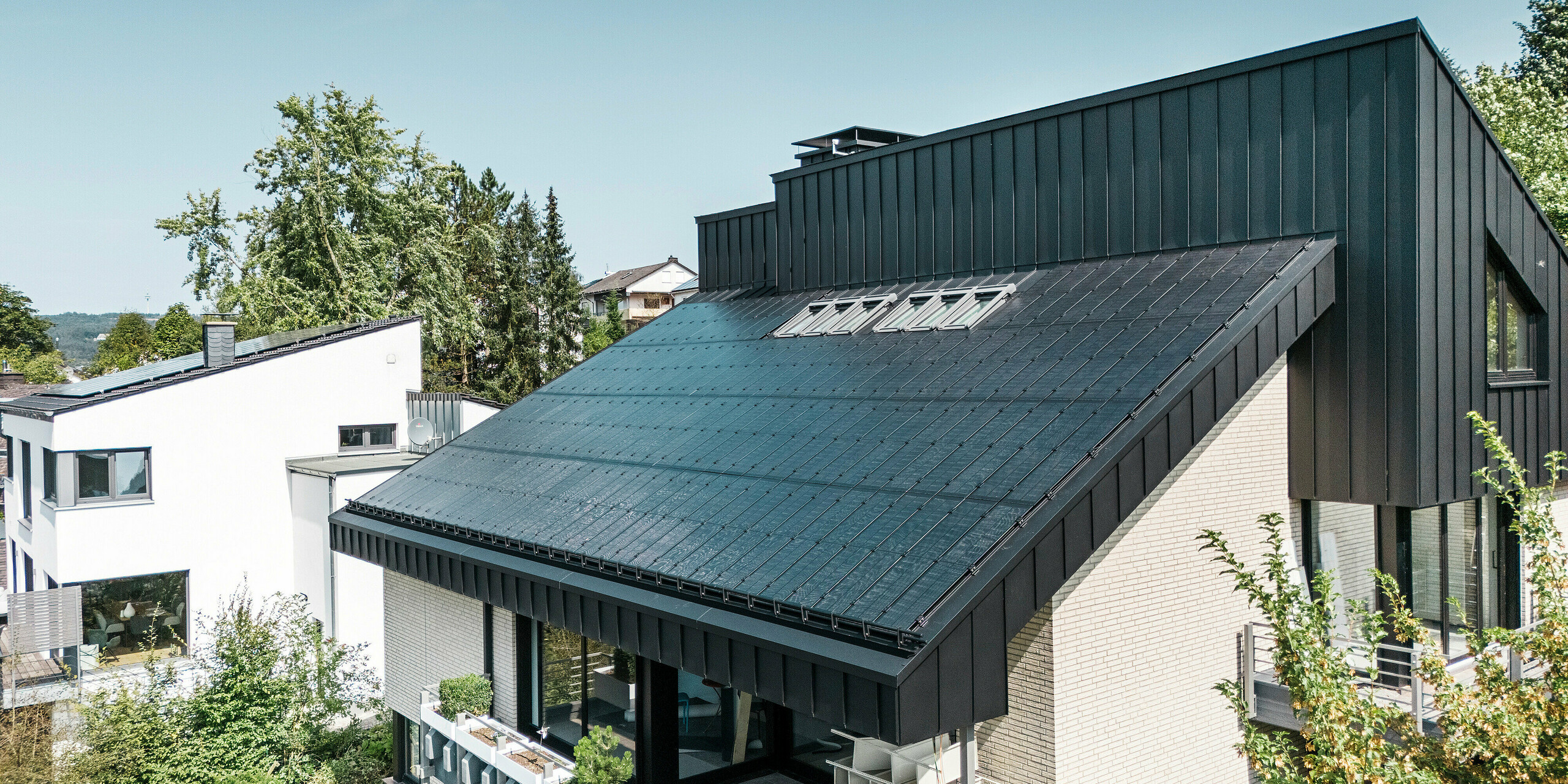 Moderne Architektur eines Einfamilienhauses in Meschede, Deutschland, mit einem PREFALZ-Aluminiumdach in P.10 Schwarz und integrierten Glas/Glas-Photovoltaikmodulen. Das versetzte Dach vereint zeitloses Design mit nachhaltiger Photovoltaik-Technologie. Die innovativen PREFALZ-Solarmodule liefern eine Leistung von 18 kWp. Die rahmenlosen Module und die elegante Aluminium-Dach- und Fassadenverkleidung bilden ein ästhetisches und funktionales Ganzes, das sich harmonisch in die grüne Wohnumgebung einfügt.